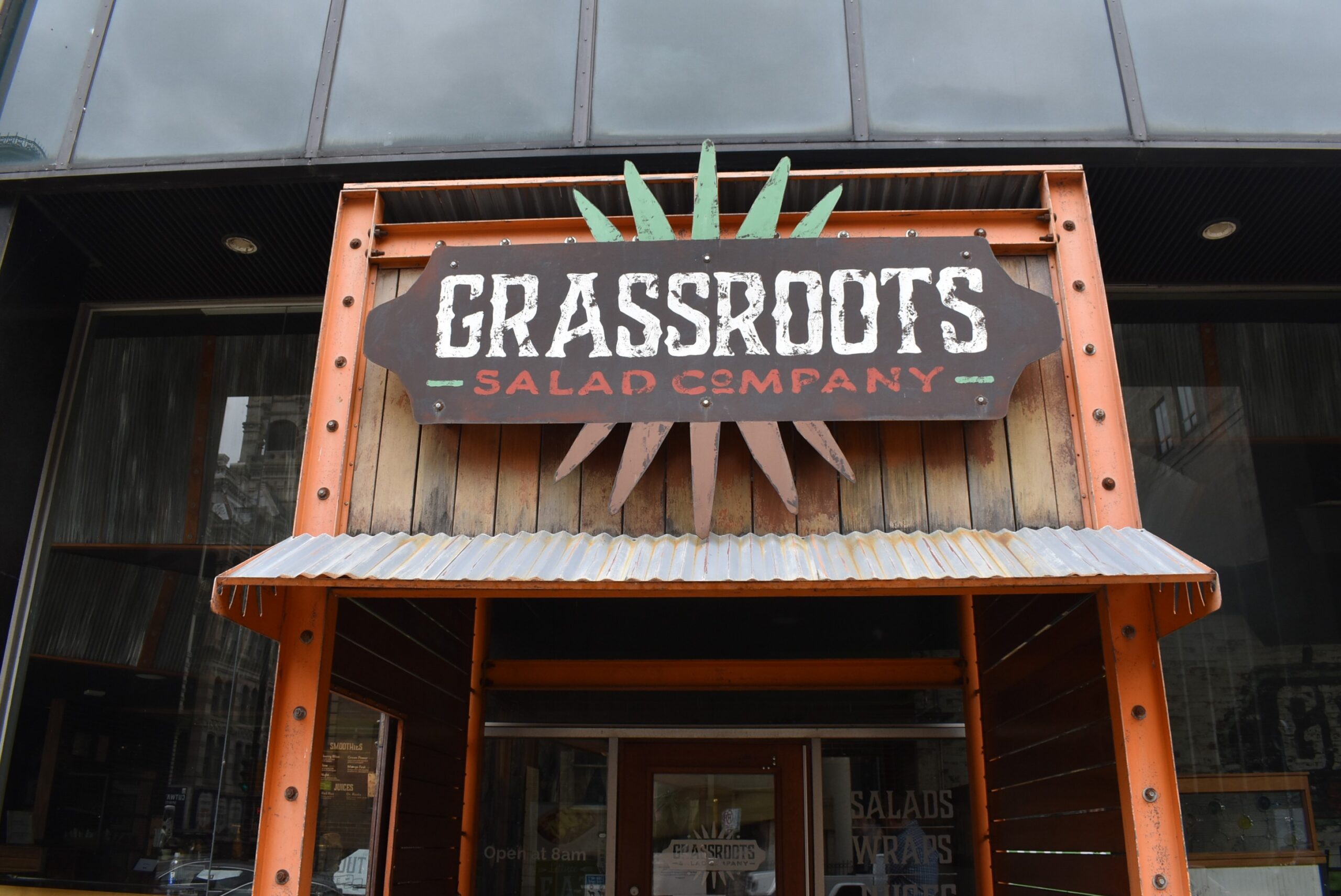 The Grassroots Salad Company storefront sign hangs above an entrance with orange trim and a corrugated metal awning.